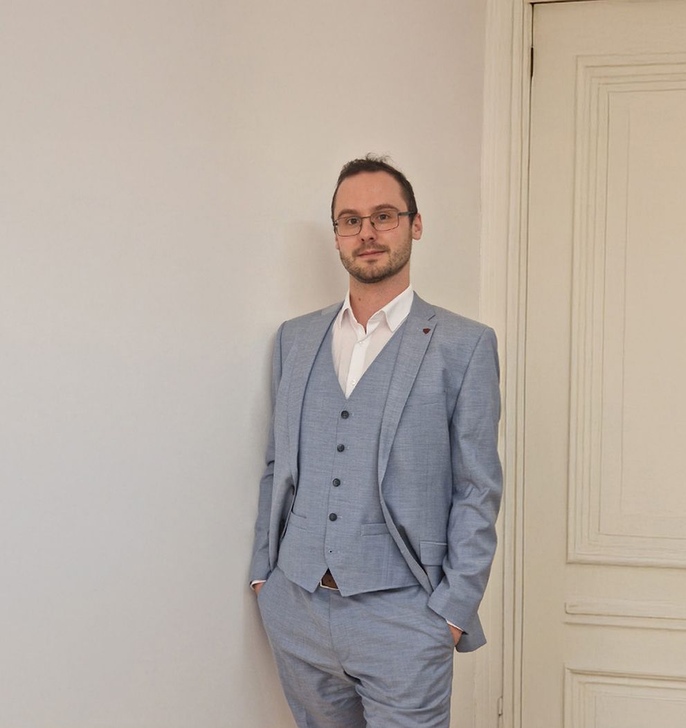 Porträtaufnahme von Mag. Peter Lippitz in einem hellblauen dreiteiligen Anzug, der selbstbewusst vor einer weißen Wand neben einer klassischen Zimmertür steht